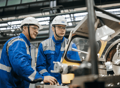 Two workers in blue safety coveralls and white hard hats operating machinery in an industrial machine shop.