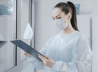 Medical professional in a mask and protective gown reviewing a patient chart in a clinical office.